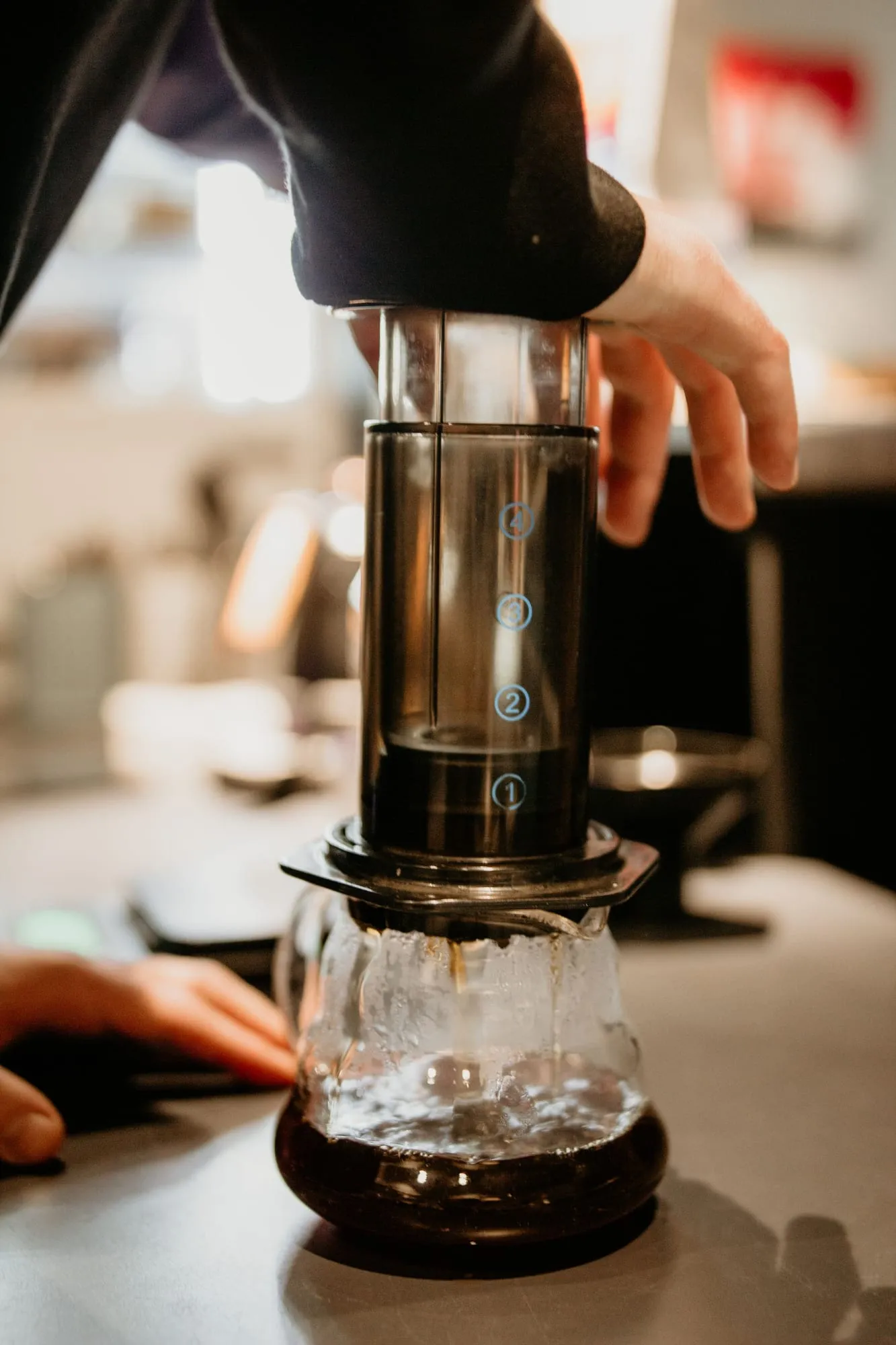 Mână care apasă pistonul unui AeroPress așezat pe un servant de sticlă